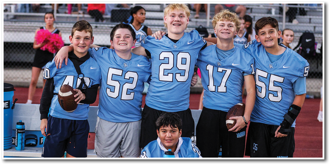 Five children in blue sports jerseys stand together on a track field with one child kneeling in front holding a water bottle, and two children holding footballs. Description generated by AI