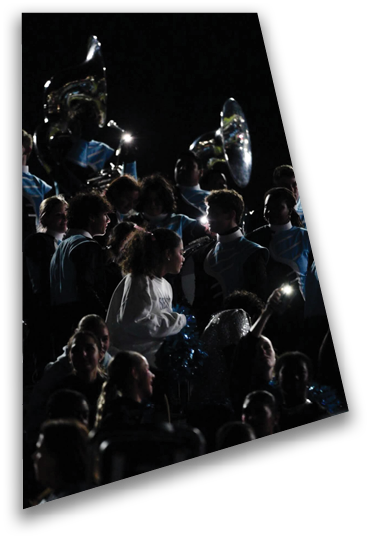 A person wearing a light-colored long-sleeve shirt is holding blue pom-poms in a dark setting with other people partially visible in the background. Description generated by AI