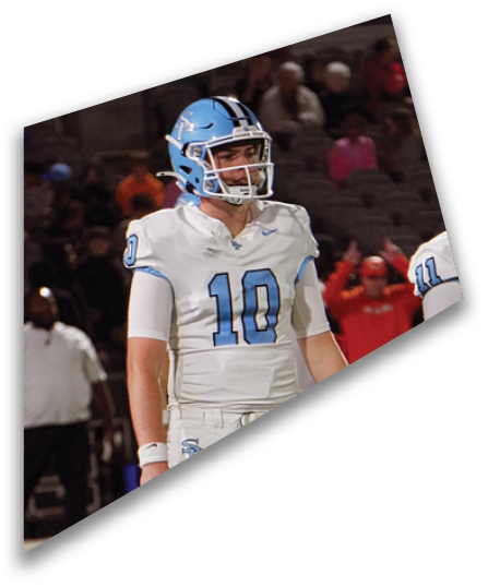 A person wearing a white football uniform with the number 10 and a light blue helmet stands on a field with blurred spectators in the background. Description generated by AI