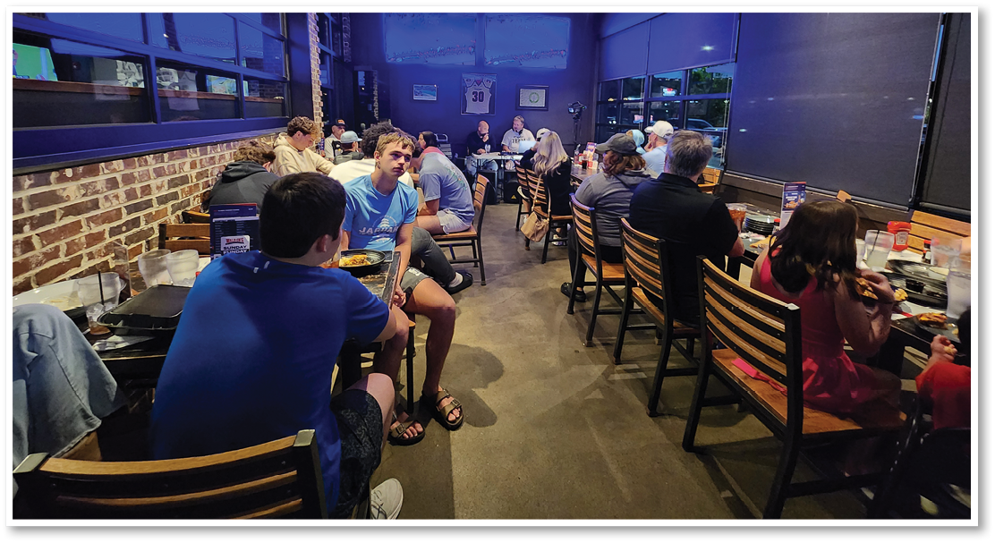 People sitting and talking at tables in a dimly lit casual restaurant with brick walls and large windows. Description generated by AI
