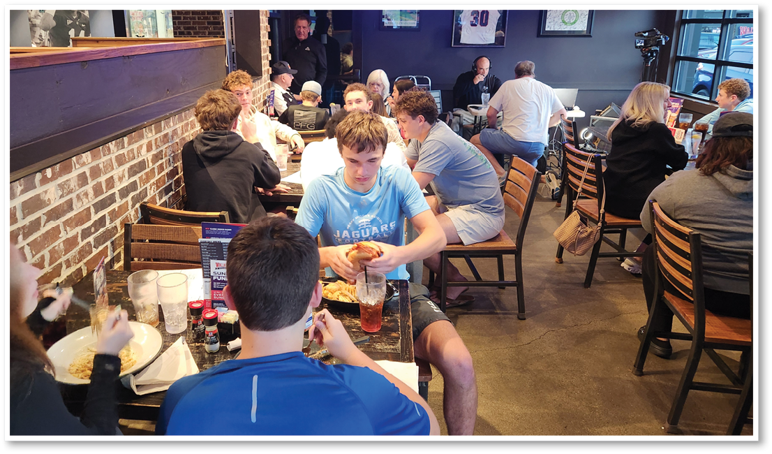 People are sitting and eating at wooden tables in a casual restaurant with brick walls and framed sports memorabilia. Description generated by AI