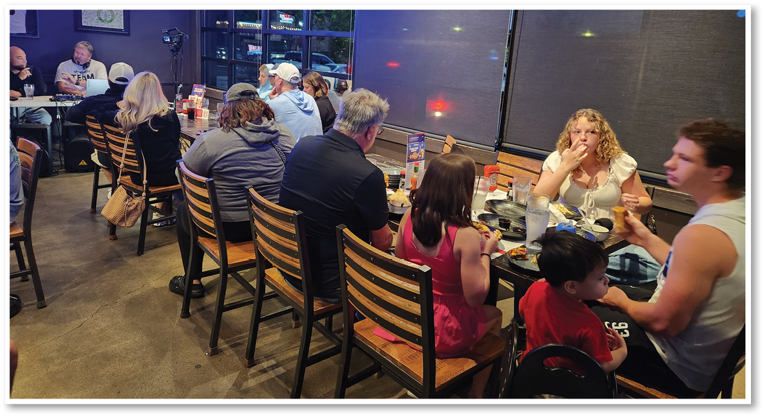 People are sitting and eating together at a restaurant with wooden chairs and tables, some engaged in conversation while others focus on their food. Description generated by AI