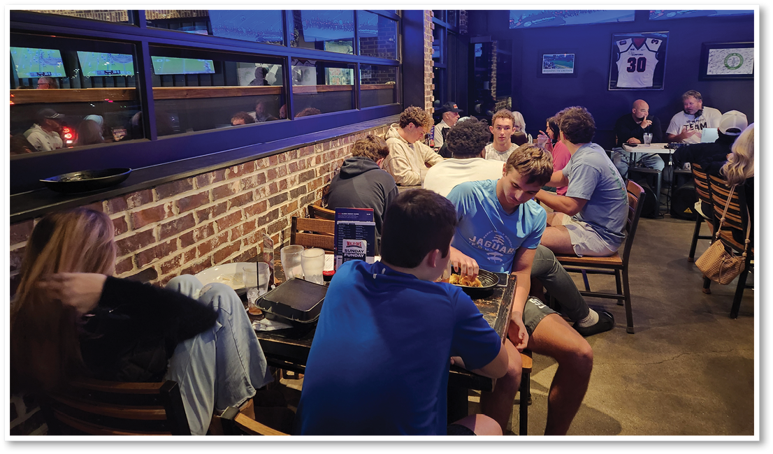 People are sitting and socializing at tables in a dimly lit casual restaurant or bar with brick walls and sports memorabilia on the walls. Description generated by AI