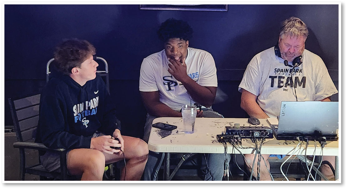 Three people wearing Spain Park team shirts are seated around a table with electronic equipment and a laptop, engaged in conversation or thought. Description generated by AI