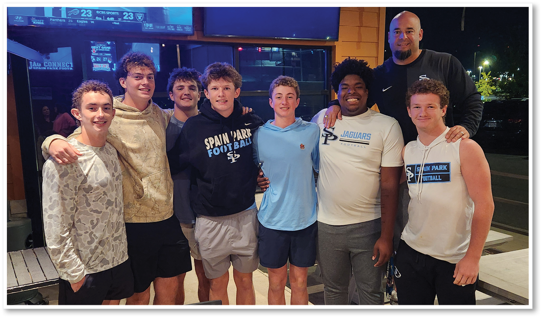A group of seven people wearing casual sportswear stand closely together outdoors at night, smiling and posing for the photo. Description generated by AI