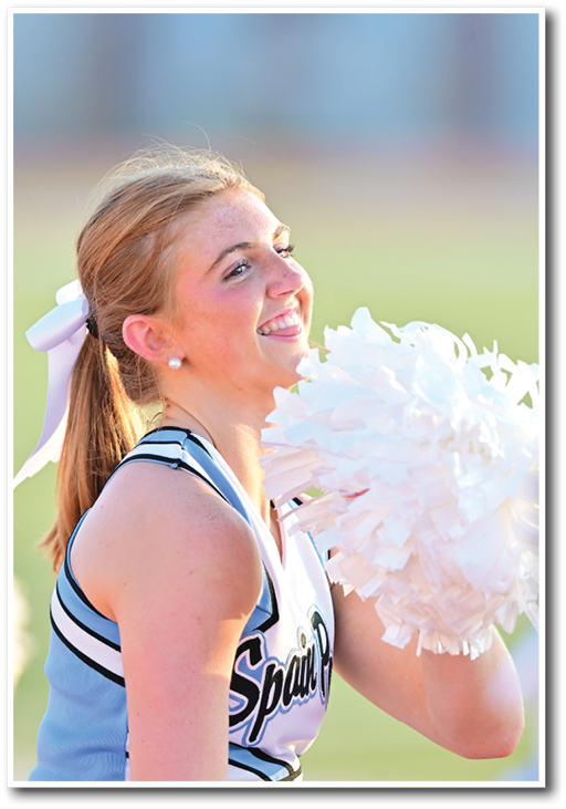 A person in a cheerleading uniform with a ponytail and a white bow is smiling and holding white pom-poms outdoors. Description generated by AI
