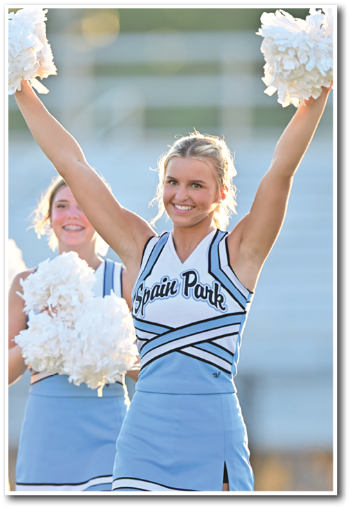 Two adults in cheerleading uniforms with “Spain Park" on the front are smiling and holding white pom-poms, with one raising both arms in the air. Description generated by AI