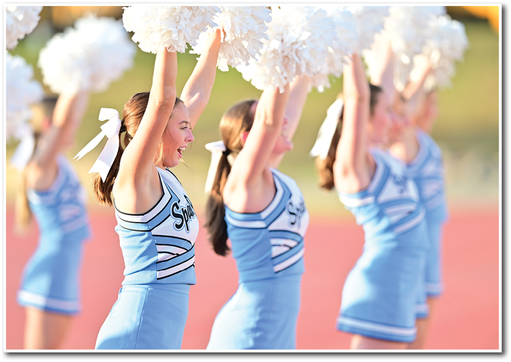 A group of adults in blue cheerleading uniforms with white bows in their hair are enthusiastically raising white pom-poms outdoors on a sports field. Description generated by AI