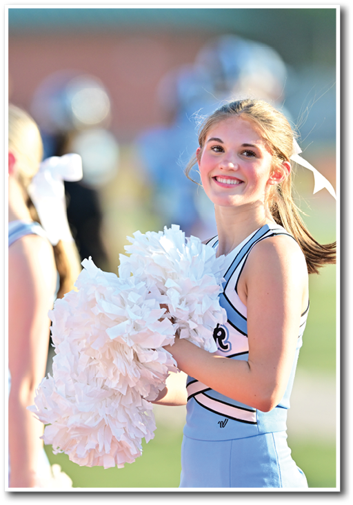 Two people in cheerleading uniforms hold white pom-poms with one smiling brightly while standing outdoors. Description generated by AI