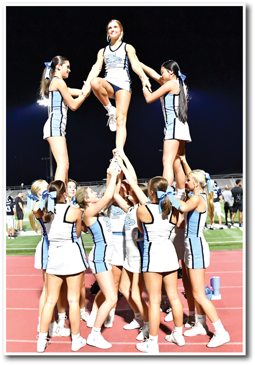 A group of people in matching cheerleading uniforms are performing a stunt on a track field at night with one person elevated above the others. Description generated by AI