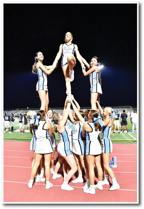 Three people in cheerleading uniforms are performing a stunt outdoors at night with one person lifted in the center holding hands with the others. Description generated by AI