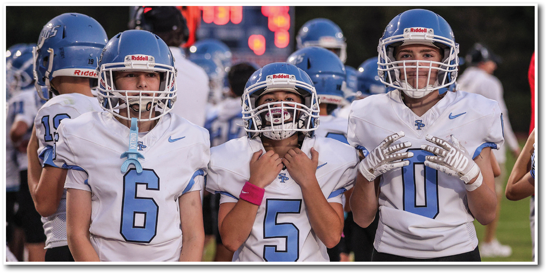 Three people wearing white and blue football uniforms and helmets stand side by side on a field with other players and a scoreboard in the background. Description generated by AI