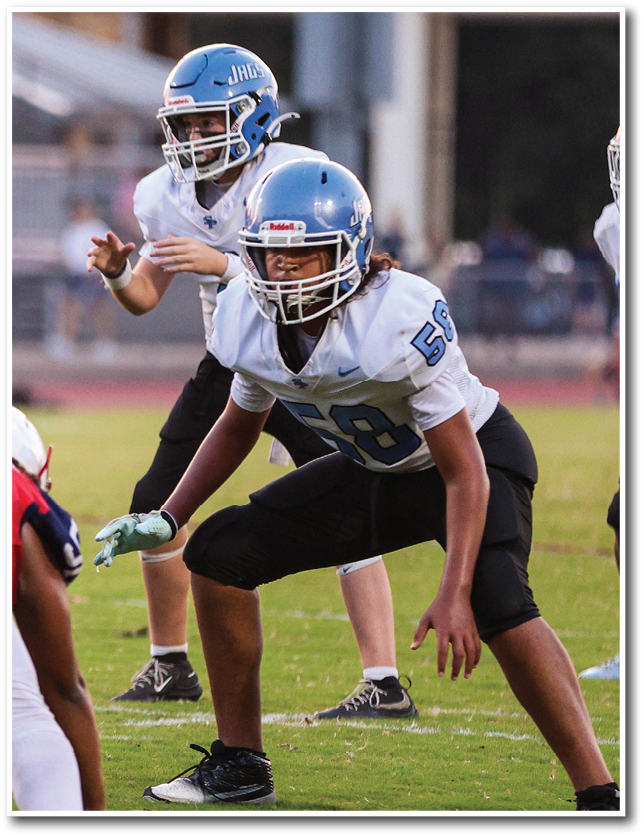 Two people wearing football helmets and uniforms are positioned on a grassy field, appearing ready to engage in a play. Description generated by AI