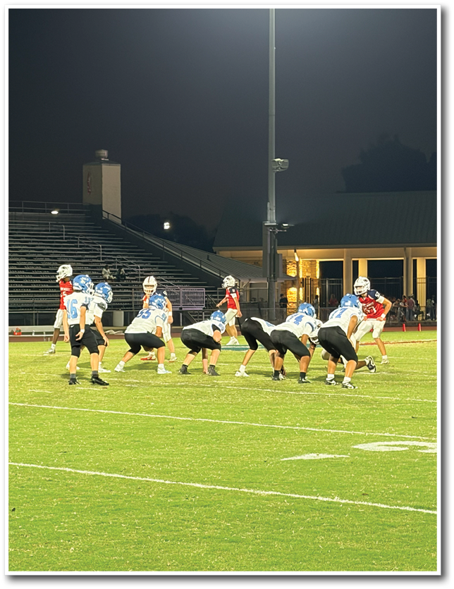 A nighttime outdoor football game shows players in blue and white uniforms lined up on the field facing players in red and white uniforms under bright stadium lights. Description generated by AI