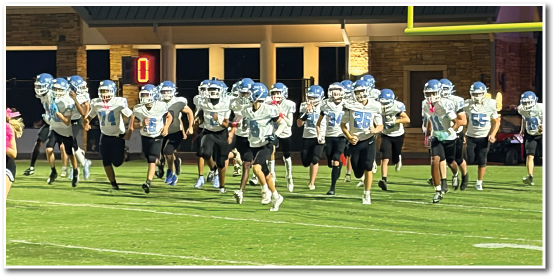 A group of people wearing helmets and sports uniforms are running on a football field under bright lights near a goalpost. Description generated by AI