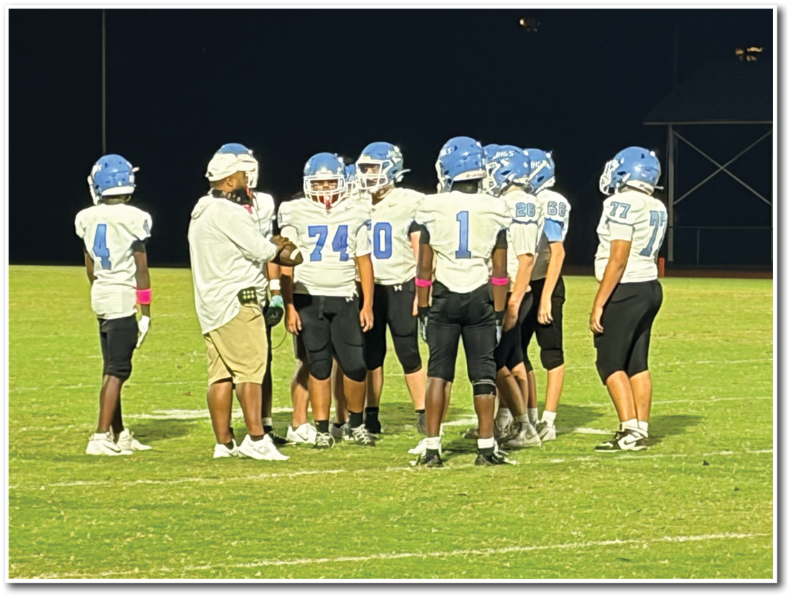 A group of people wearing football uniforms and helmets are standing on a grass field at night, with one person in casual clothing appearing to give instructions. Description generated by AI