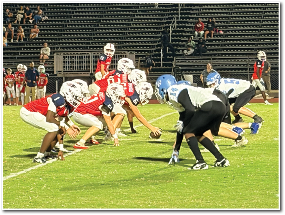 Two opposing football teams in red and white and black and blue uniforms are lined up facing each other on a grassy field with empty bleachers in the background. Description generated by AI