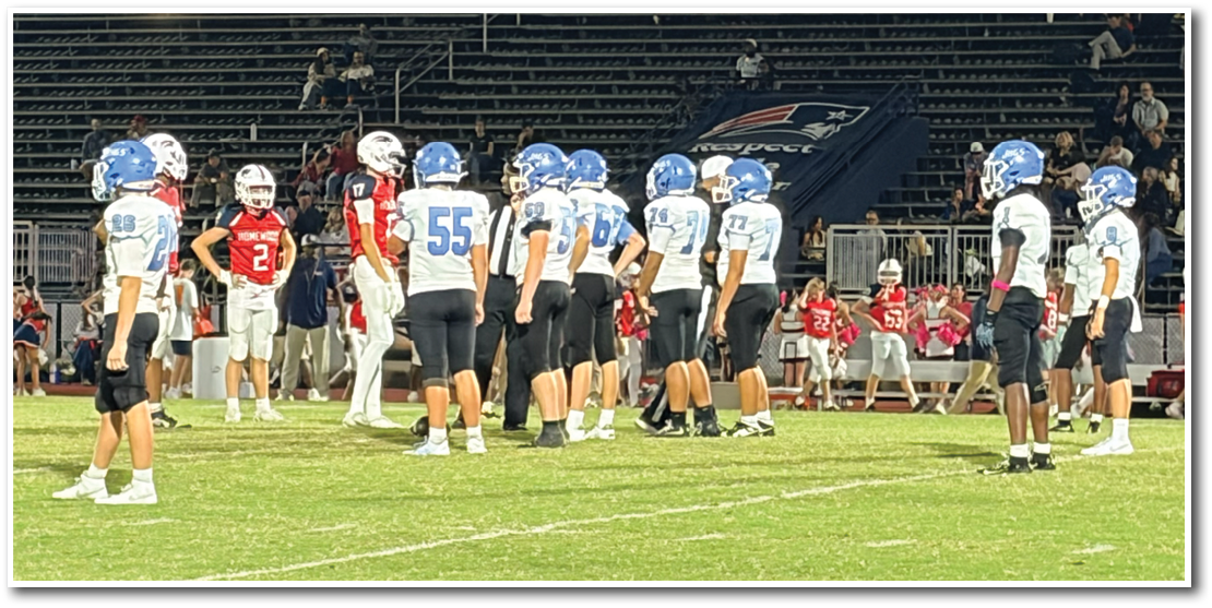 Players from two football teams in helmets and uniforms stand on a field facing each other with empty bleachers and a few spectators in the background. Description generated by AI