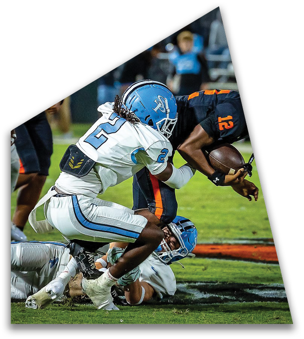 A person in a dark uniform holding a football is being tackled by two people in white and blue football uniforms on a grassy field. Description generated by AI