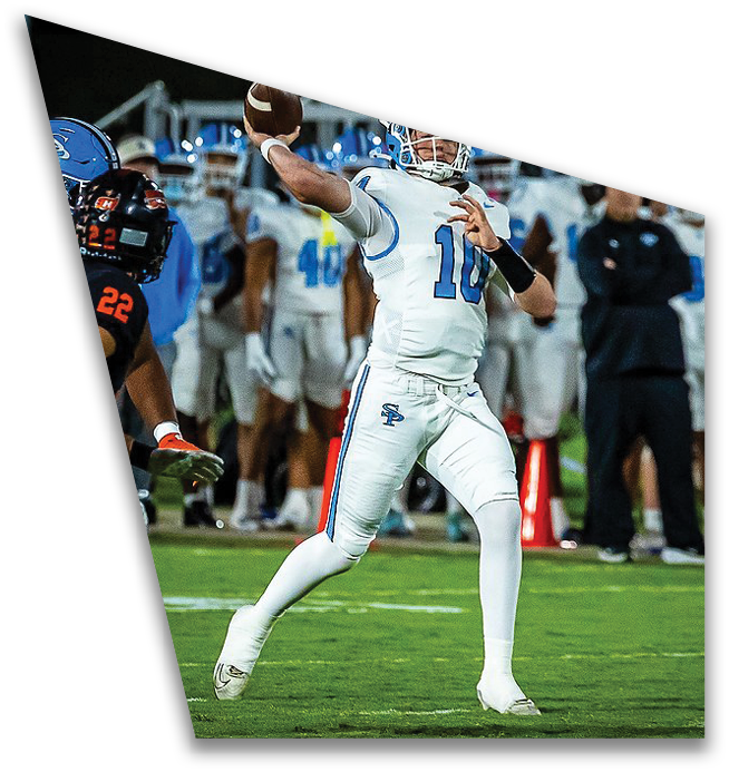 A person in a white football uniform is preparing to throw a football while another person in a black and orange uniform approaches on a grassy field with a crowd in the background. Description generated by AI