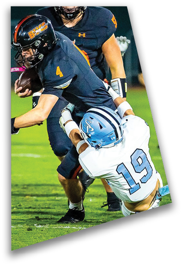 A person in a dark football uniform is running with the ball while being tackled by another person in a white football uniform on a grassy field during a game. Description generated by AI