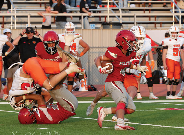 Four people wearing black football uniforms with “VISTA" on the front are standing together in a stadium with a crowd in the background, two of them holding helmets raised above their heads. Description generated by AI