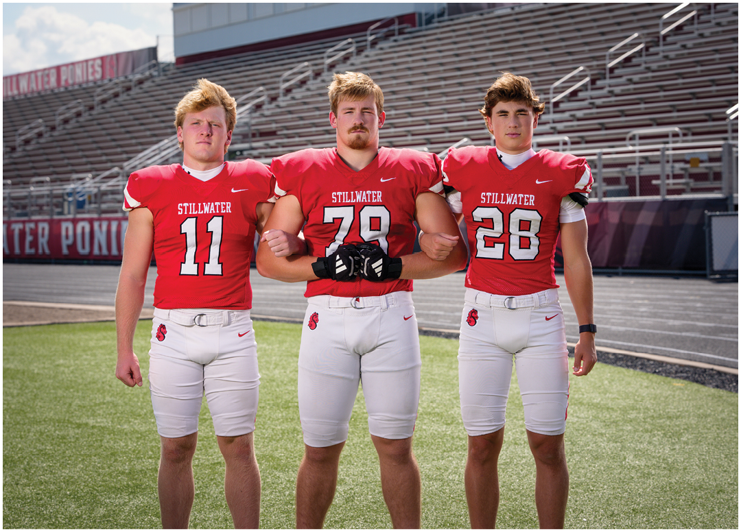 Three adults wearing red football jerseys with the word “Stillwater" and white pants stand arm in arm on a football field with empty bleachers in the background. Description generated by AI