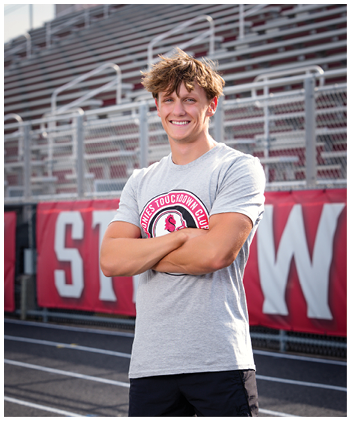 A person with tousled hair wearing a gray t-shirt with a sports logo stands smiling with arms crossed on a track field in front of bleachers and a red banner. Description generated by AI