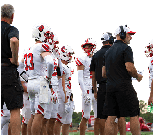 A group of people wearing white football uniforms and helmets stand on a field alongside coaches dressed in black shirts and shorts. Description generated by AI