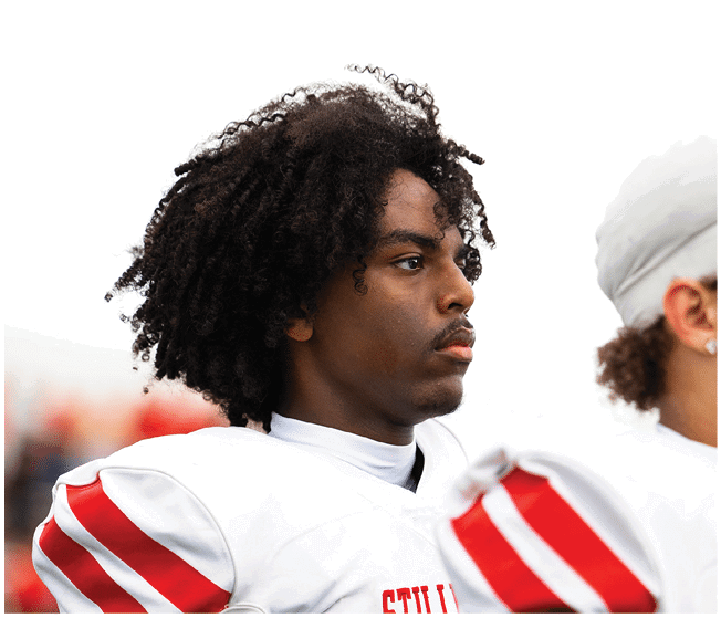 A person with curly hair wearing a white sports jersey looks intently to the side against a blurred outdoor background. Description generated by AI