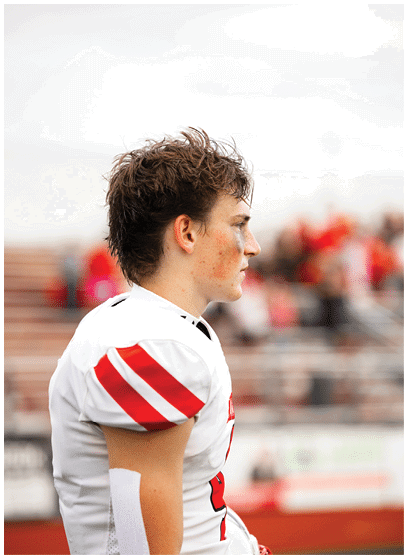 A person with short, tousled hair wearing a white sports jersey with red stripes on the sleeves is standing in profile on a sports field with blurred spectators in the background. Description generated by AI