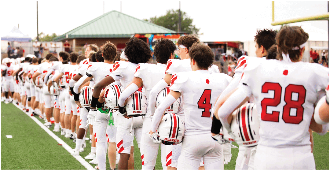 A line of people wearing white football uniforms with red numbers stand side by side on a field, holding their helmets and facing away from the camera. Description generated by AI