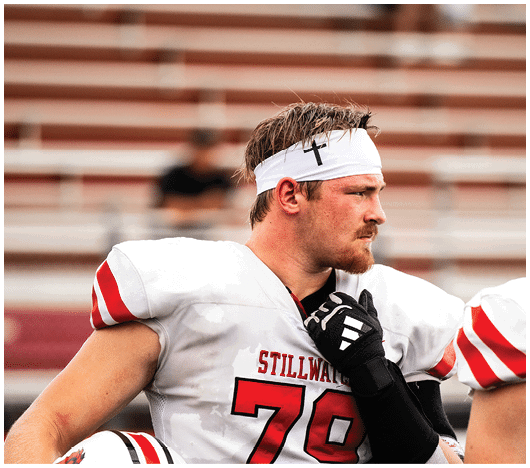 A person wearing a white sports jersey with red stripes and a white headband with a black cross is looking to the side while holding a black glove near their chest. Description generated by AI