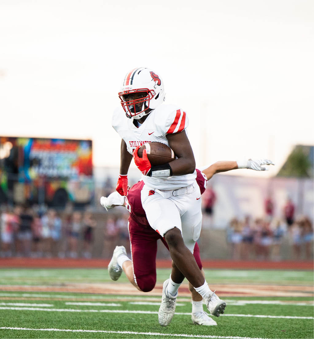 A person in a white football uniform with red stripes is running with a football while another player in a maroon uniform reaches out behind them on a field. Description generated by AI