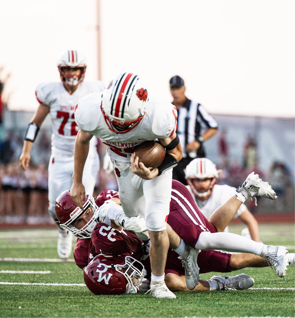 A person in white football gear is running with the ball while two people in maroon football uniforms tackle them on a grassy field. Description generated by AI
