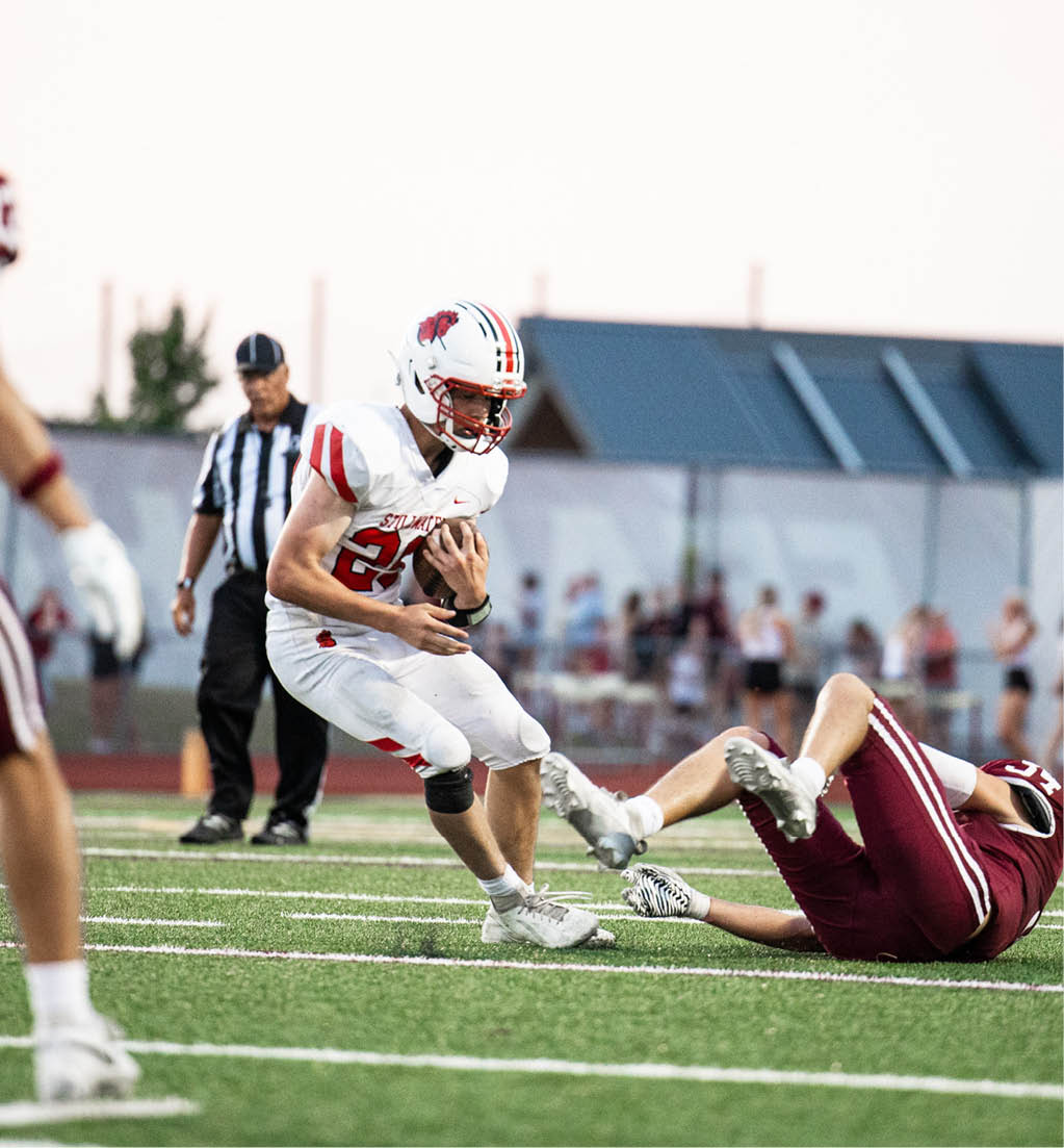 A person in a white football uniform is running with the ball while another person in a maroon uniform is falling on the ground during a game on a grassy field with a referee and spectators in the background. Description generated by AI