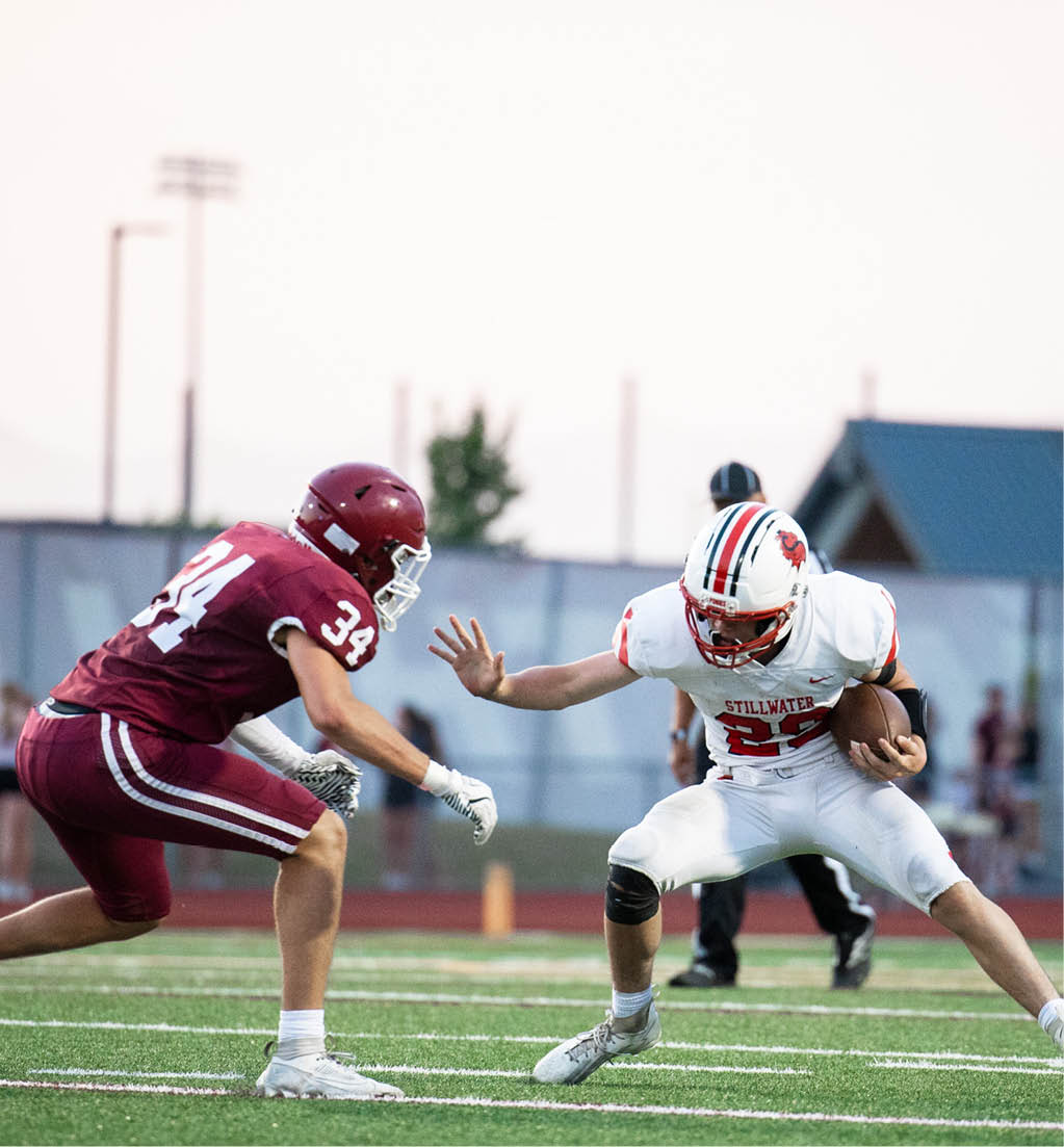 Two people in football uniforms are engaged in a play on a field, with one person in a maroon uniform crouching defensively and the other in a white uniform holding the ball and preparing to move. Description generated by AI