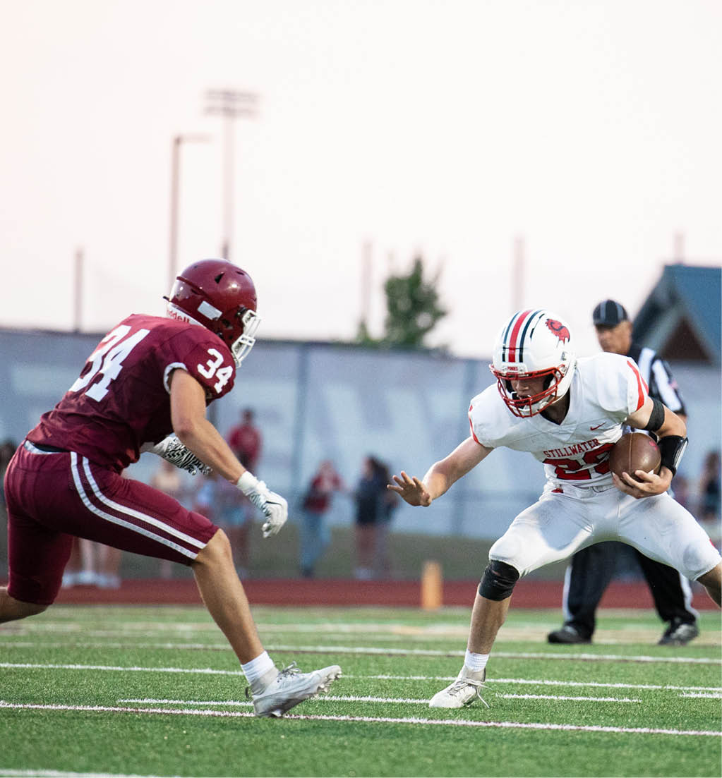 Two people in football uniforms are engaged in a play on a field, with one person in a maroon uniform running toward another person in a white uniform who is in a defensive stance. Description generated by AI