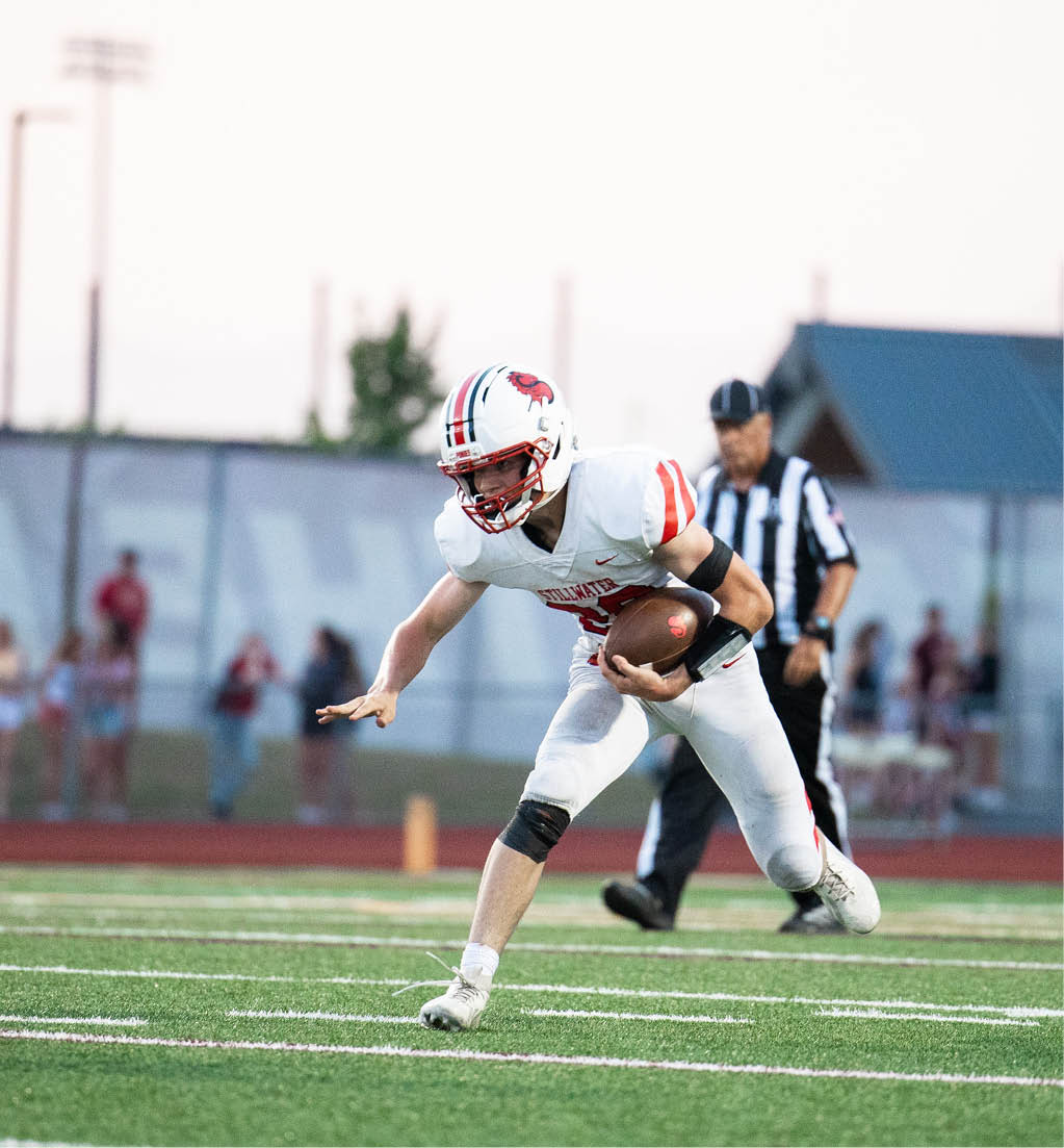 An adult in a white football uniform is running on a field while holding a football with a referee and blurred spectators in the background. Description generated by AI