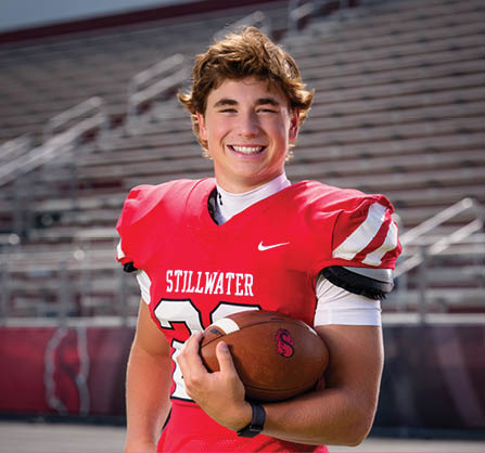 A person wearing a red football jersey with the word Stillwater and holding a football is standing on a field with empty bleachers in the background. Description generated by AI
