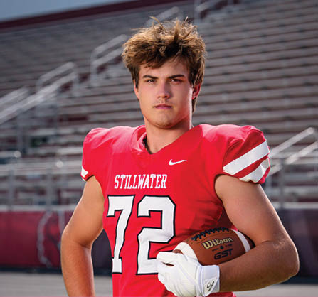 A person with tousled hair wearing a red football jersey with the number 72 and white gloves is holding a football on a sports field with empty bleachers in the background. Description generated by AI