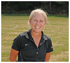 A person with light hair tied back is smiling and wearing a black collared sports shirt while sitting outdoors on a grassy field. Description generated by AI