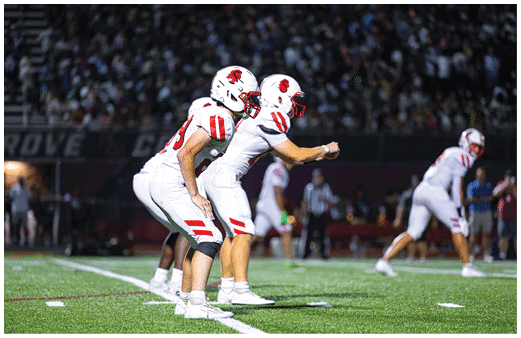 Two people in white sports uniforms stand on a football field, with one person clapping and the other facing away. Description generated by AI