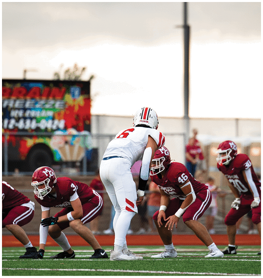 A group of people wearing white football uniforms with red and black accents are sitting and standing closely together on a sports field. Description generated by AI