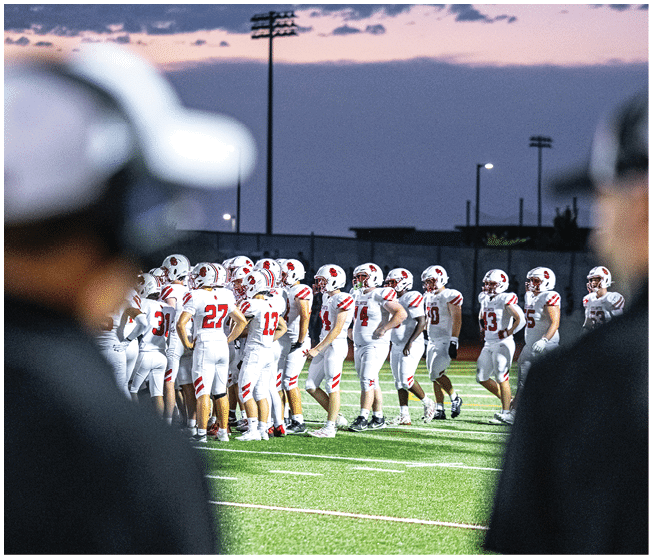 A group of people wearing white football uniforms with red and black accents are sitting and standing closely together on a sports field. Description generated by AI