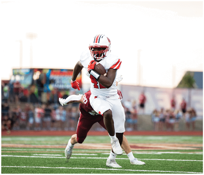 A group of people wearing white football uniforms with red and black accents are sitting and standing closely together on a sports field. Description generated by AI