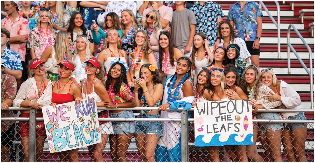 A large group of people wearing colorful Hawaiian shirts and leis are gathered in bleachers, smiling and holding signs that say “WE RUN THIS" and "WIPE OUT THE LEAFS." Description generated by AI