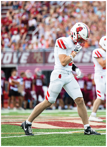 A person in a white football uniform with red accents is holding a football and appears to be running during a game. Description generated by AI