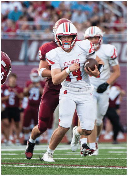 A person in a white football uniform with red accents is holding a football and appears to be running during a game. Description generated by AI