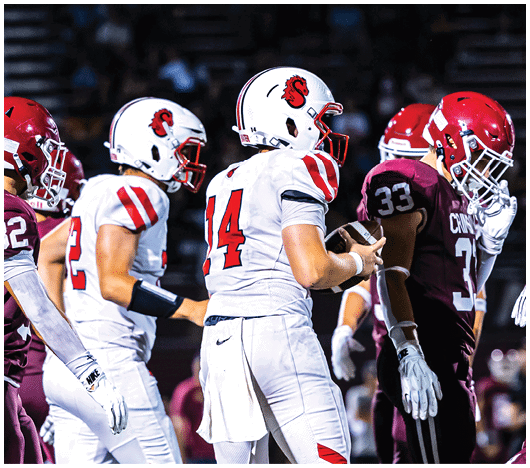 Two people wearing white football helmets with red faceguards and a red lion logo are focused and looking ahead. Description generated by AI